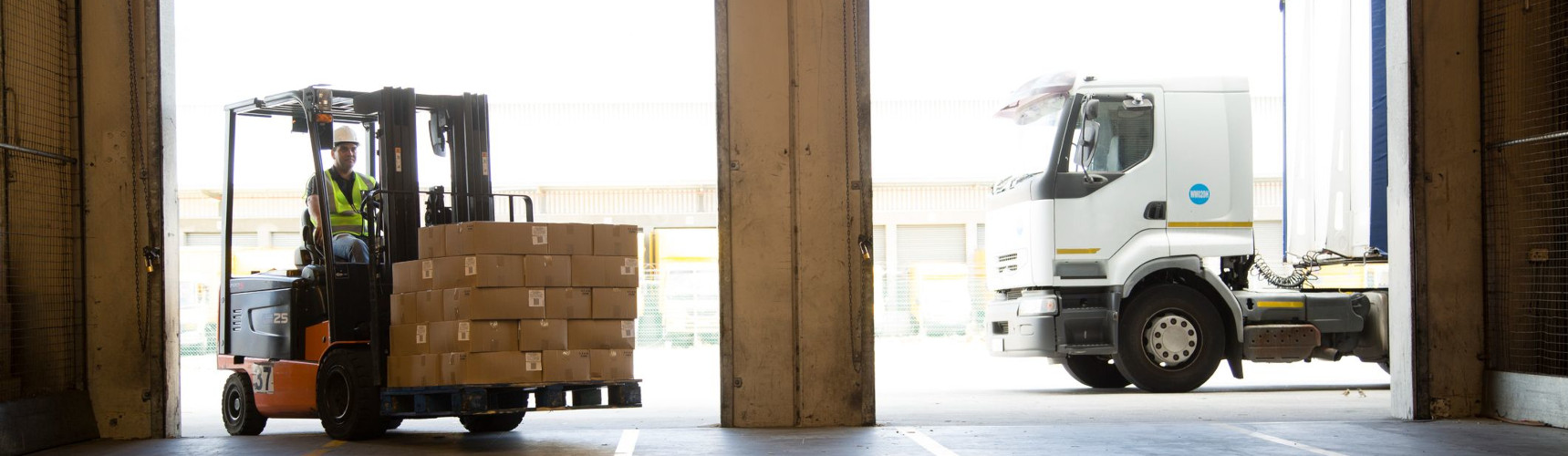 Gestão de supply chain: uma máquina carrega várias caixas de entregas dentro de uma armazém, ao lado da porta há um caminhão